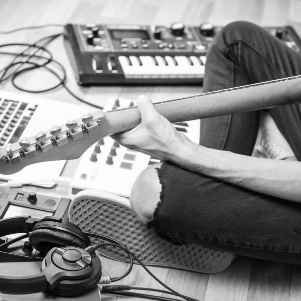 Male guitarist playing electric guitar at modern home studio or rehearsal room. Young man producing music with electronic effects processors, synthesizer and laptop